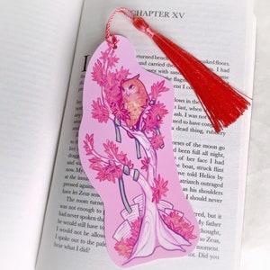 Può includere: Un segnalibro rosa con un fiocco rosso. Il segnalibro presenta un'illustrazione stilizzata di un gufo appollaiato su un ramo con fiori rosa. Il gufo è marrone e bianco con occhi gialli. Il segnalibro ha la forma di una lacrima.