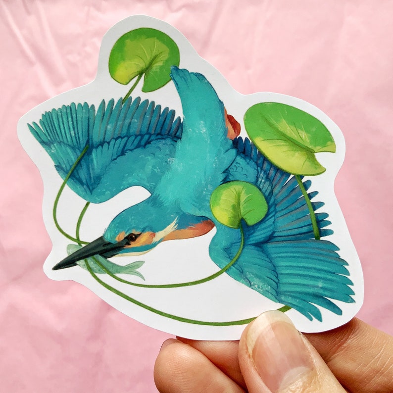 Puede incluir: Un mart&iacute;n pescador azul con nen&uacute;fares verdes, un pez peque&ntilde;o en su pico y un fondo blanco.