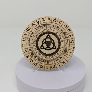 Puede incluir: Una rueda de cifrado circular de madera con un diseño de triskel en el centro. La rueda tiene un anillo exterior giratorio con letras del alfabeto y un anillo interior fijo con runas y símbolos.
