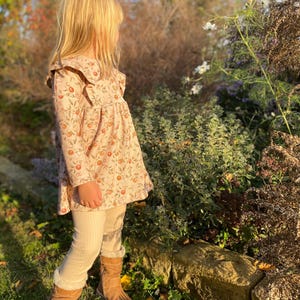 以下が含まれることがあります： フリル襟のついた長袖の花柄ワンピースとクリーム色のレギンスを着用した少女。彼女は茶色のブーツを履いて、緑の芝生と花のある庭に立っています。