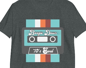 Puede incluir: Camiseta gris oscuro con un diseño de cinta de cassette retro. La cinta es turquesa y gris, con las palabras "Boogie Down" y "70's Soul" impresas. El diseño incluye rayas verticales en turquesa, blanco y naranja.