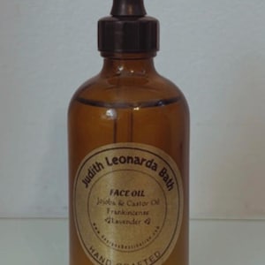 May include: A brown glass bottle of face oil with a dropper. The label reads "Judith Leonarda Bath FACE OIL Jojoba & Castor Oil Frankincense Lavender HAND CRAFTED". The bottle is filled with a clear liquid.