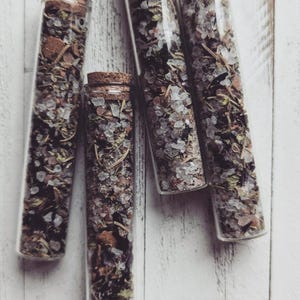 May include: Five clear glass vials filled with dried herbs, crystals, and other natural elements. Each vial is sealed with a cork stopper. The contents are a mix of green, brown, and white, creating a rustic and earthy aesthetic. The vials are arranged on a white wooden surface.