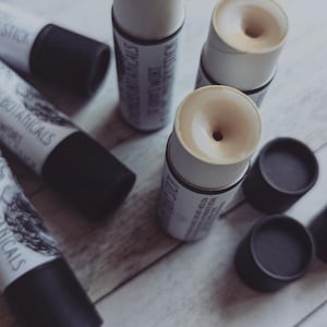 May include: Five tubes of lip balm with a white label and black text. The label reads "Botanicals" and "Lip Treatment". The tubes are on a light wood surface.