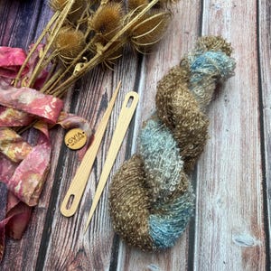 May include: A skein of brown and blue yarn with two wooden needles, dried flowers, and a pink ribbon. The yarn is a mix of brown and blue fibers. The wooden needles are long and slender. The ribbon is pink and red.