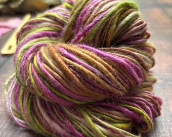 May include: A close-up of a skein of yarn with a blend of colors. The yarn features shades of green, purple, brown, and tan. The yarn is tightly wound, showing the texture and color variations. Ideal for knitting and crochet projects.