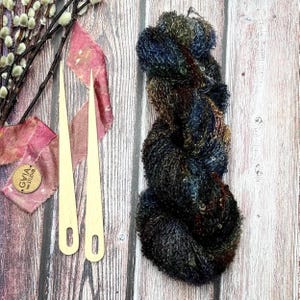 Könnte beinhalten: Ein Strang aus strukturiertem Garn in dunklen Blau-, Braun- und Schwarztönen mit eingearbeiteten Pailletten, begleitet von zwei hellen Webnadeln aus Holz. Das Garn hat ein flauschiges, mehrfarbiges Aussehen mit dezentem metallischem Glanz.