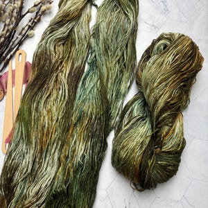 May include: Close-up of hand-dyed yarn in shades of green, brown, and gold. The yarn is arranged in skeins and a ball, showcasing the color variations. The yarn is likely for knitting or crochet projects.
