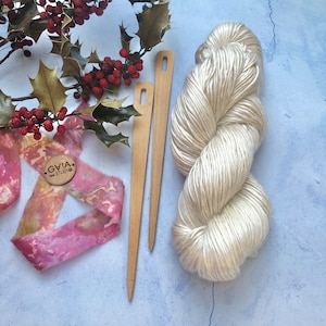 Peut inclure: Un écheveau de fil couleur crème, deux aiguilles à tricoter en bois clair, une branche de houx aux baies rouges et un morceau de tissu à motifs rose et jaune avec une étiquette ronde en bois marquée GVIA FIBRE STUDIO.