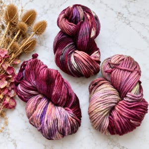 Könnte beinhalten: Drei Stränge aus dickem, handgefärbtem Garn in variierenden Schattierungen von tiefem Magenta, Pflaume, zartem Rosa und Creme. Das Garn weist eine grobe, gedrehte Textur mit fließenden Farbübergängen in jedem Strang auf.