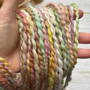 May include: A close-up of a skein of colorful yarn. The yarn is a mix of pastel colors, including pink, yellow, green, and cream, with a silver metallic thread woven throughout. The yarn is thick and textured, perfect for knitting or crocheting.
