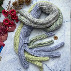 May include: A soft, knitted scarf in shades of light green and blue. The scarf is folded and arranged on a marble surface. A small, round tag with the text "GYIA" is visible in the lower right corner of the image.