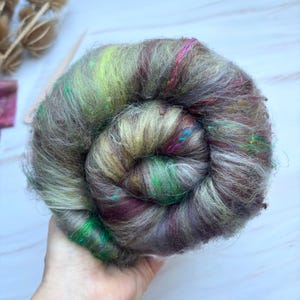 May include: A hand holds a tightly wound ball of colorful yarn. The yarn is a mix of greens, browns, purples, and pinks, with a soft, textured appearance. The yarn is likely used for knitting or other fiber crafts.