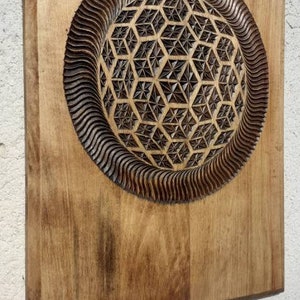 K&ouml;nnte beinhalten: Ein Wandbehang aus Holz mit einem geschnitzten geometrischen Muster in der Mitte. Das Muster ist dunkelbraun und von einem hellbraunen Holzrahmen umgeben.