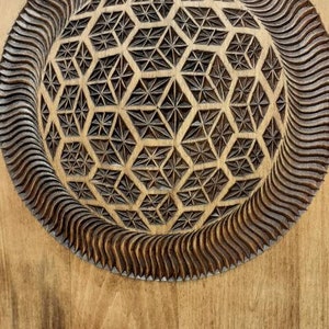 K&ouml;nnte beinhalten: Ein Wandbehang aus Holz mit einem geschnitzten geometrischen Muster in der Mitte. Das Muster ist von einem wellenf&ouml;rmigen Rand umgeben.