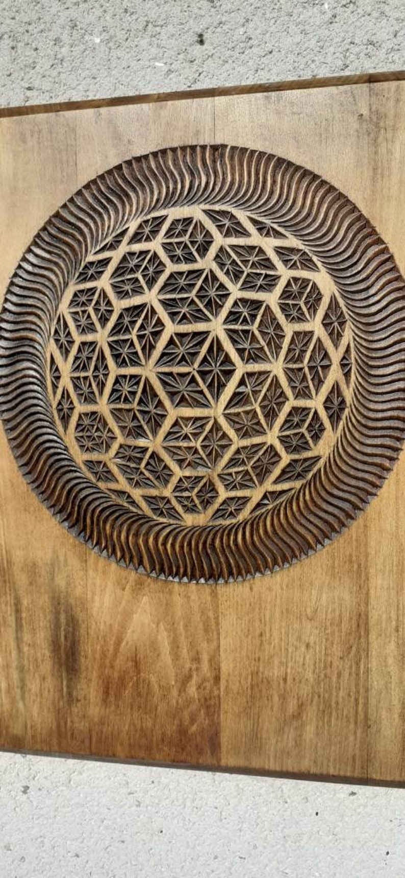 K&ouml;nnte beinhalten: Ein Wandbehang aus Holz mit einem geschnitzten geometrischen Muster in der Mitte. Das Muster ist von einem wellenf&ouml;rmigen Rand umgeben.
