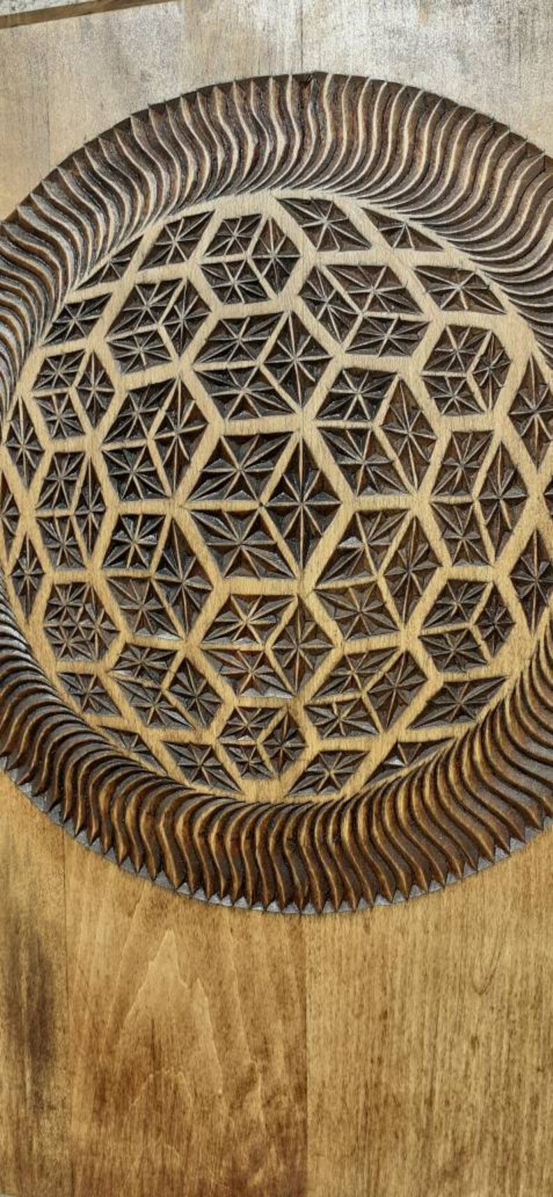 K&ouml;nnte beinhalten: Eine Holzplakette mit einem geschnitzten geometrischen Muster in der Mitte. Das Muster ist ein Kreis mit einem sich wiederholenden Muster aus Dreiecken und Sternen. Die Plakette ist hellbraun und hat eine glatte Oberfl&auml;che.