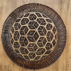 K&ouml;nnte beinhalten: Ein Wandbehang aus Holz mit einem geschnitzten geometrischen Muster in der Mitte. Das Muster ist eine Kugel mit einem sich wiederholenden Muster aus Sechsecken und Dreiecken. Die Kugel ist von einem wellenf&ouml;rmigen Rand umgeben.