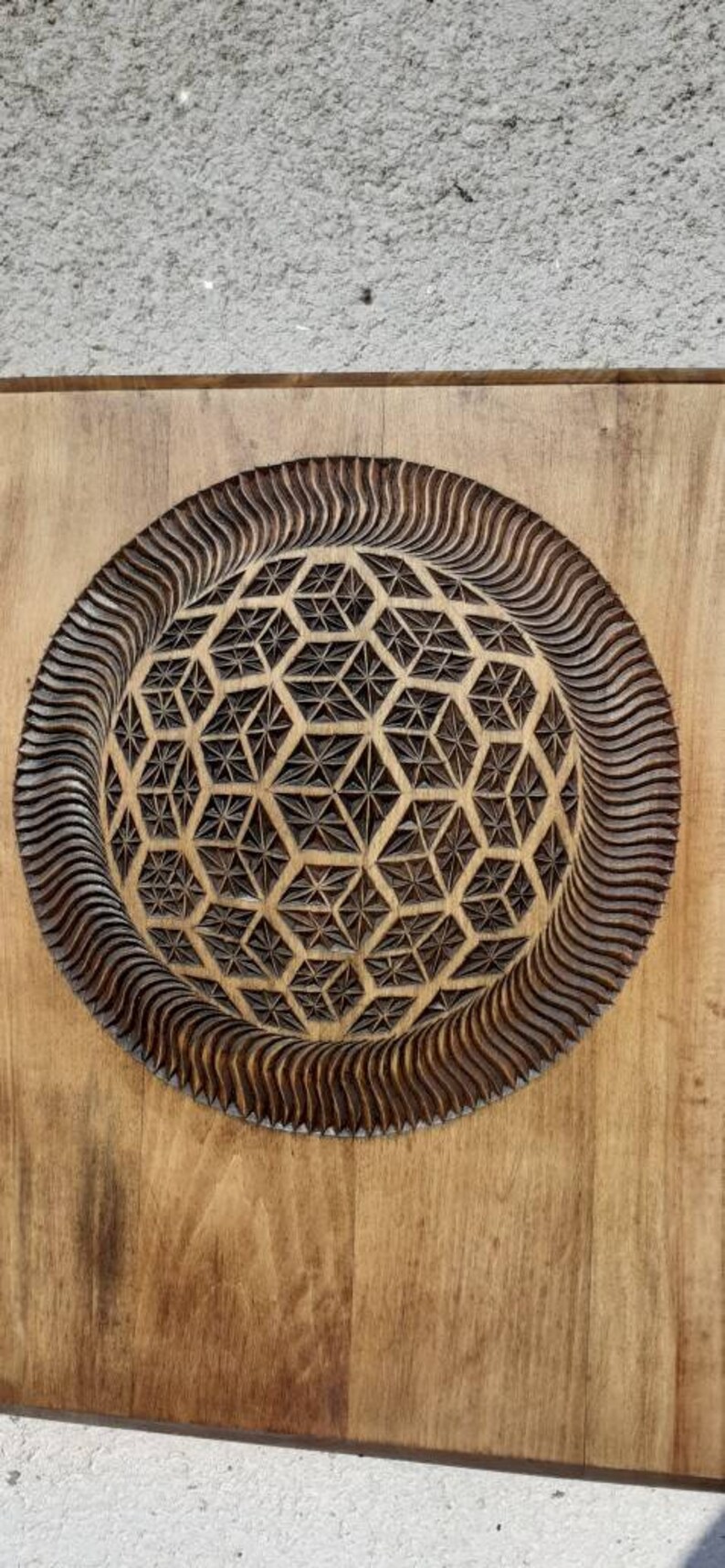 K&ouml;nnte beinhalten: Ein Wandbehang aus Holz mit einem geschnitzten geometrischen Muster in der Mitte. Das Muster ist eine Kugel mit einem sich wiederholenden Muster aus Sechsecken und Dreiecken. Die Kugel ist von einem wellenf&ouml;rmigen Rand umgeben.