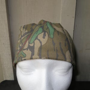 May include: A camouflage print beanie hat in shades of green, brown, and tan. The hat is made of a soft fabric and is designed to fit snugly on the head. The hat is a practical accessory for outdoor activities.