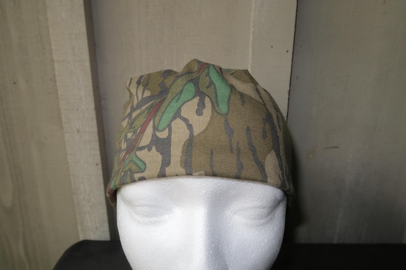 May include: A camouflage print beanie hat in shades of green, brown, and tan. The hat is made of a soft fabric and is designed to fit snugly on the head. The hat is a practical accessory for outdoor activities.