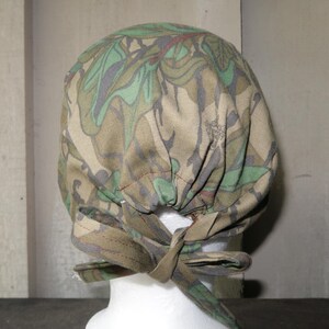 May include: A camouflage patterned scrub cap with a tie closure. The cap features a mix of green, brown, and tan colors. The tie is made of the same camouflage fabric.