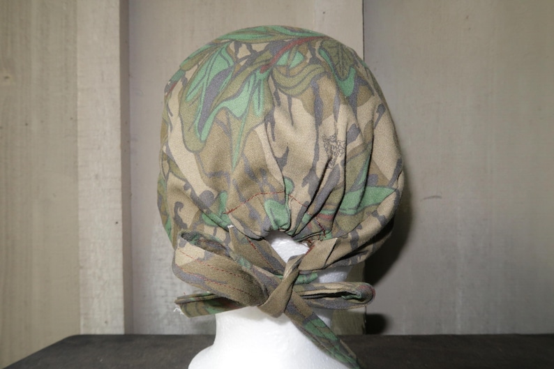 May include: A camouflage patterned scrub cap with a tie closure. The cap features a mix of green, brown, and tan colors. The tie is made of the same camouflage fabric.