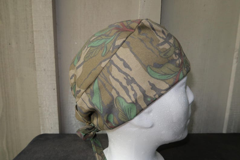 May include: A camouflage print head covering, featuring a pattern of green leaves and brown and gray shapes. The head covering is tied at the back. The fabric appears to be cotton or a similar material.
