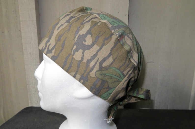 May include: Camouflage pattern head covering. The fabric features a blend of brown, green, and gray hues, creating a woodland camouflage design. The head covering is tied at the back.