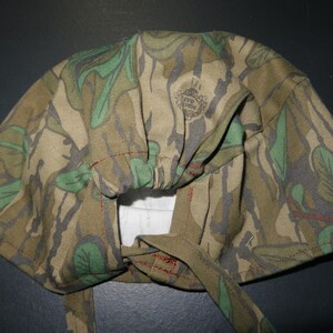 May include: A camouflage-patterned head covering with a tie closure. The fabric features a mix of green, brown, and gray leaf and branch designs. The head covering has a gathered section and is stitched with red thread.