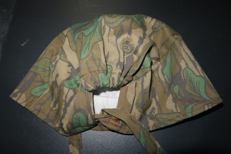 May include: A camouflage-patterned head covering with a tie closure. The fabric features a mix of green, brown, and gray leaf and branch designs. The head covering has a gathered section and is stitched with red thread.