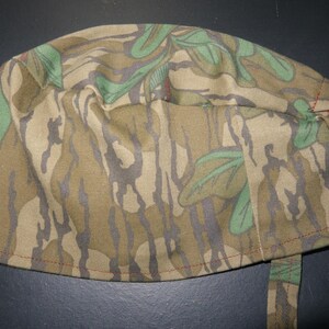 May include: A camouflage print scrub cap in shades of brown, green, and gray. The cap features a tie closure and is made of a soft, flexible fabric. The seams are stitched with red thread.