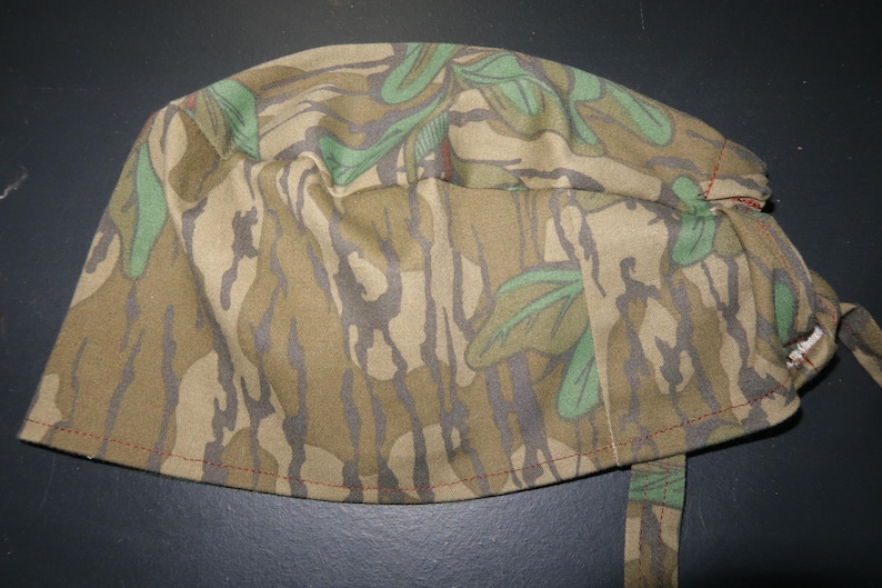 May include: A camouflage print scrub cap in shades of brown, green, and gray. The cap features a tie closure and is made of a soft, flexible fabric. The seams are stitched with red thread.