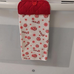 May include: A red and white Christmas-themed kitchen towel hanging on an oven. The towel features a pattern of red candy canes, snowflakes, and candies on a white background. The top portion is a quilted red oven mitt with a button.