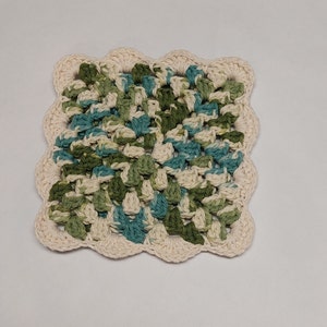 May include: A crocheted square dishcloth in shades of green, blue, and white. The dishcloth has a scalloped edge.
