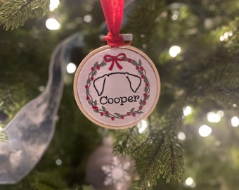 May include: A white, round, embroidered Christmas ornament with a red ribbon and a green holly wreath. The ornament features a white dog silhouette with the name "Cooper" embroidered in black.