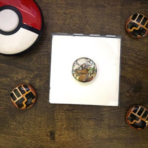 May include: A red, white, and black Poké Ball sits next to a white square with a round, illustrated button. Three additional round buttons with a black and gold design are also present. The items are on a dark brown wooden surface.