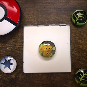 May include: A collection of decorative magnets and a Pokeball on a wooden surface. The magnets feature various designs, including a star, a leaf, and a cartoon character. The Pokeball is red and white.
