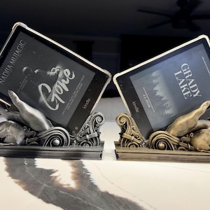 May include: Two e-reader stands featuring sculpted hands emerging from ornate, swirling bases. One stand is finished in a metallic silver, holding a Kindle displaying the book Gone by Nadija Mujagic. The other is a metallic gold, holding a Kindle displaying Grady Lake.