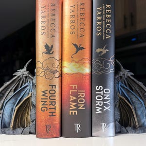 May include: Three hardcover books by Rebecca Yarros titled Fourth Wing, Iron Flame, and Onyx Storm, positioned between two dark gray dragon wing bookends. The book spines feature gold and silver text, dragon silhouettes, and cloud illustrations.