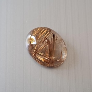 May include: Oval-shaped gemstone with a clear, translucent base. The stone features intricate, needle-like inclusions in shades of gold and brown, creating a striking visual texture. The gemstone is polished and smooth.