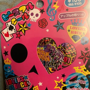 May include: A pink and black package with a skull design and the text "Lovers Rock". The package contains 70 stickers and one large sticker. The stickers are themed with skulls, stars, hearts, and music notes. The package also says "Apple scent".