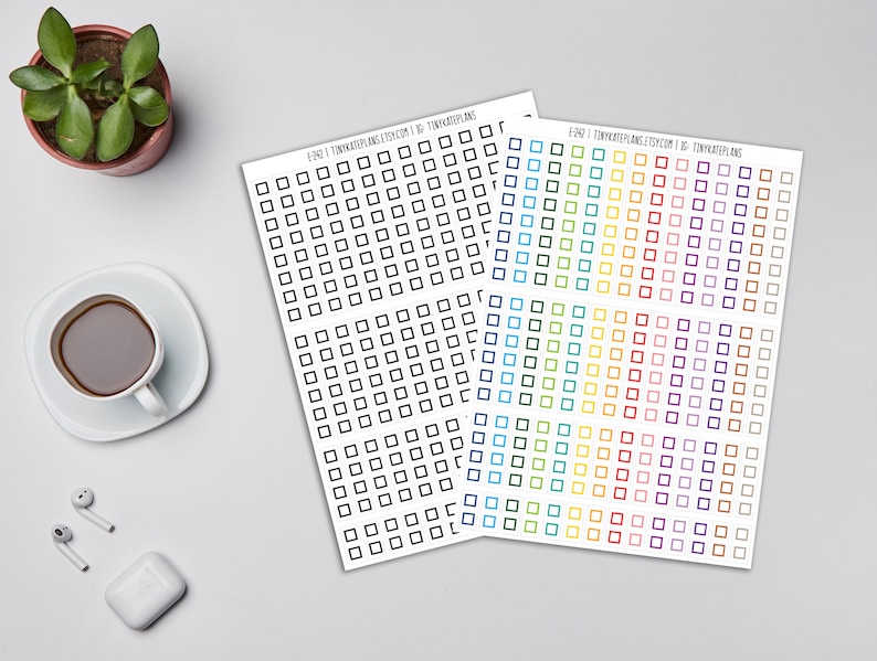 May include: Sheets of square stickers in black and rainbow colours. The stickers are arranged in a grid pattern. The text "E242 | TINYKATEPLANS.ETSY.COM | IG: TINYKATEPLANS" is printed on the top of the sheets. A cup of coffee, a small plant, and earbuds are also visible.