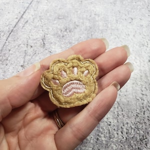 May include: A small, brown, fuzzy paw print patch with a pink embroidered paw print in the center.