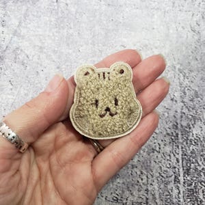 May include: A brown fuzzy bear face patch with a white outline. The bear has a simple embroidered smile.