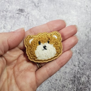 May include: A brown fuzzy bear face applique with white fur around the nose and mouth. The bear face is outlined in yellow thread.