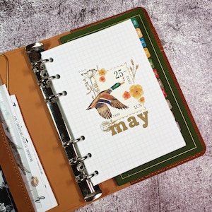 May include: A white planner with a grid pattern and a decorative sticker featuring a duck, flowers, and a stamp that says "25 - Post 1880 LCN" and "May".