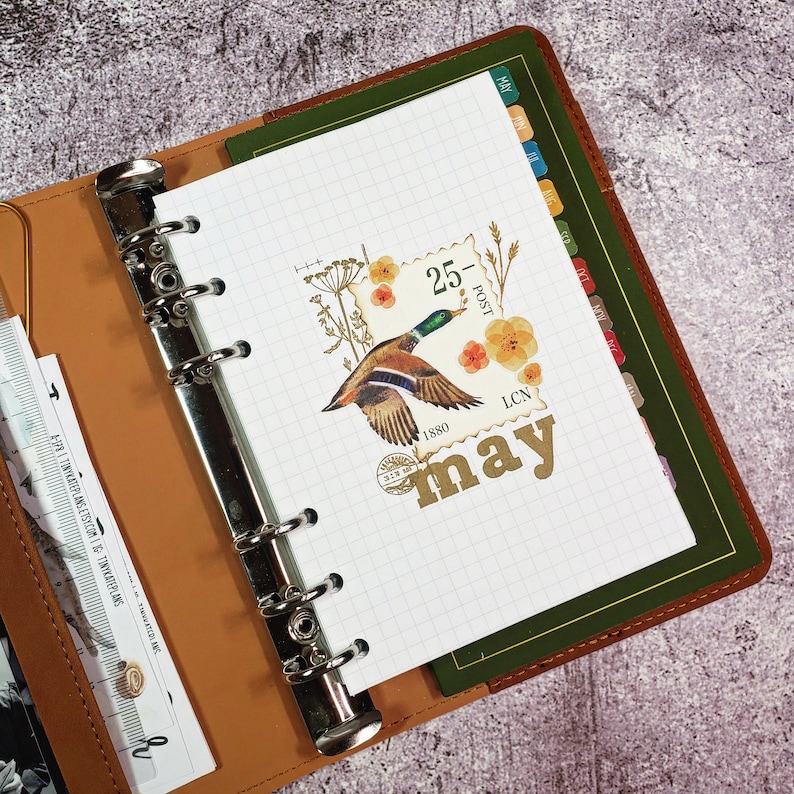 May include: A white planner with a grid pattern and a decorative sticker featuring a duck, flowers, and a stamp that says "25 - Post 1880 LCN" and "May".