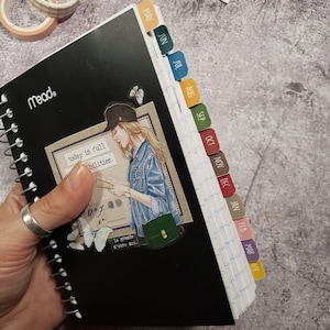 May include: A black spiral bound notebook with monthly tabs. The tabs are color coded and labeled with the months of the year. The notebook has a quote on the front that says "today is full of possibilities".