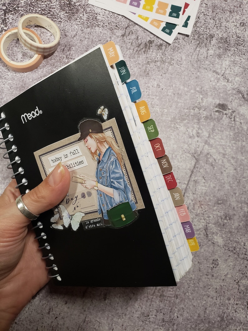 May include: A black spiral bound notebook with monthly tabs. The tabs are color coded and labeled with the months of the year. The notebook has a quote on the front that says "today is full of possibilities".